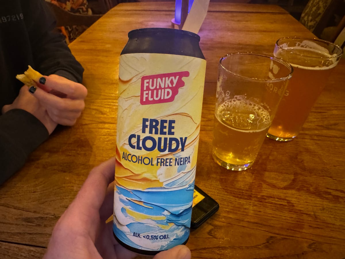 En hånd holder en dåse Funky Fluid Free Cloudy ved et træbord med ølglas i baggrunden