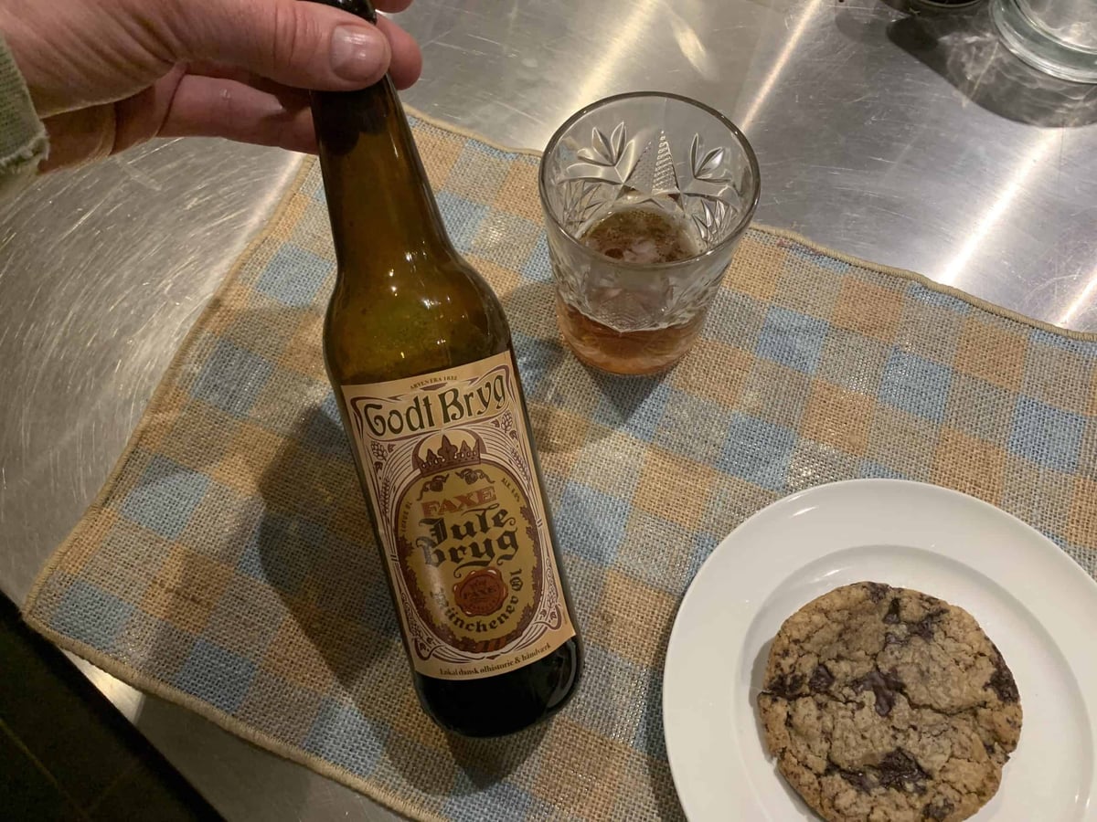 En hånd holder Godt Bryg Faxe Julebryg ved et glas ravfarvet øl og en småkage på en tallerken