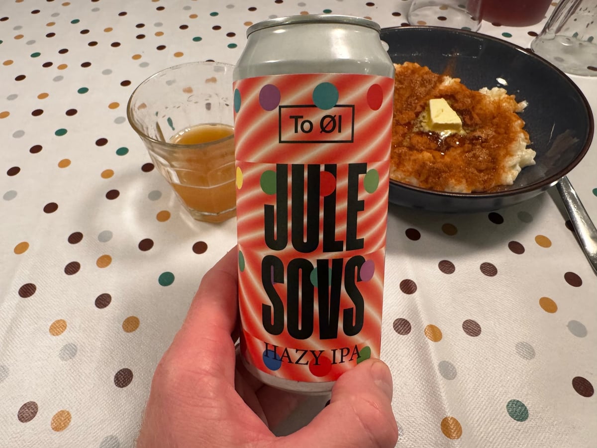 En hånd holder en dåse To Øl Jule Sovs foran et glas øl og et måltid