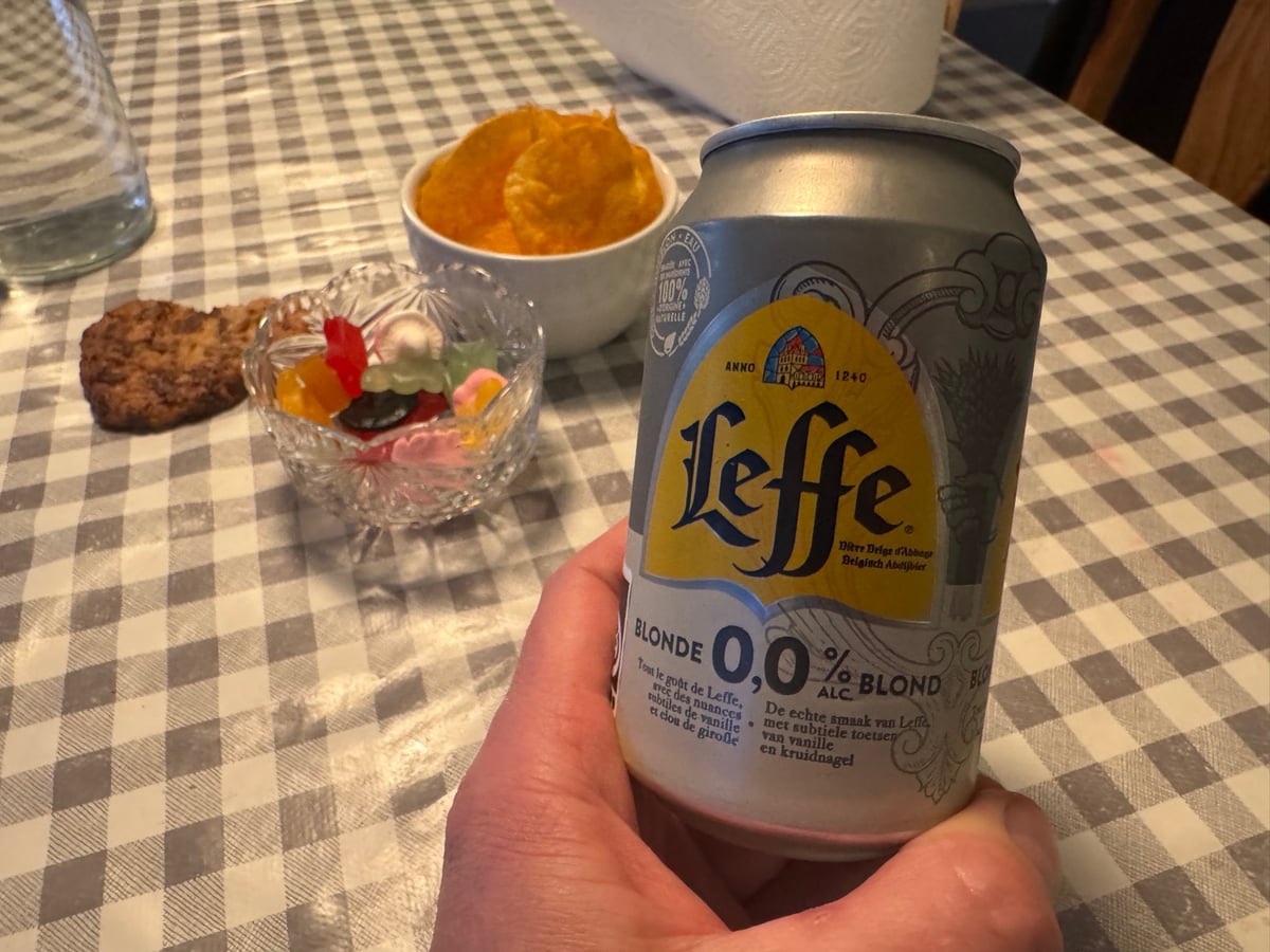 En hånd holder en dåse Leffe Blonde 0,0 % over et ternet bord med snacks i baggrunden
