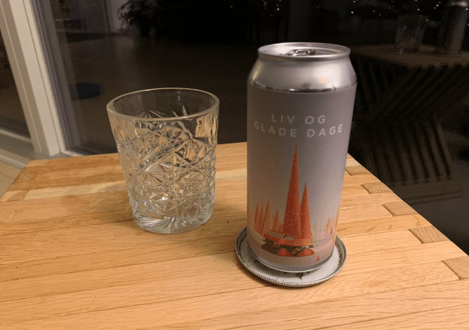 Grå dåse Liv og Glade Dage fra Ølsnedkeren med orange tegning ved siden af et tomt glas på en træflade